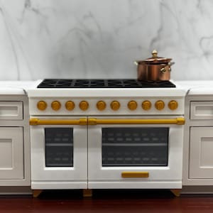 May include: A miniature white kitchen range with a black stovetop, gold-colored knobs, and gold-colored handles on two oven doors. A small copper-colored pot with a lid sits on the stovetop. The range is flanked by white cabinetry.