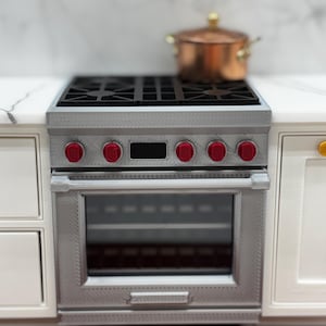 May include: A miniature silver-colored kitchen range with a black stovetop grate, six bright red control knobs, and a glass-front oven door. A small copper-colored pot with a lid sits on the stovetop burner above the oven.