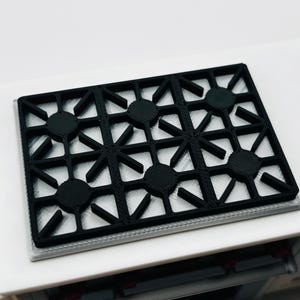 May include: A black, rectangular stovetop with six burners. The burner grates are a geometric design. The stovetop is set on a white surface, with a glimpse of a cabinet below.