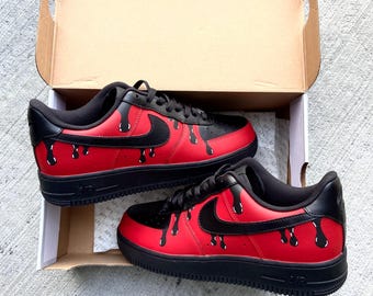Op de afbeelding: Een paar rode en zwarte sneakers met een druppelend verfdesign. De sneakers hebben een zwart swoosh-logo en een zwarte zool.