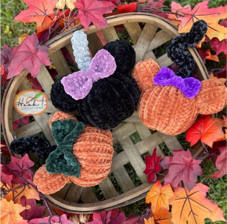 May include: Three crocheted pumpkin-shaped ornaments with different colored bows. The pumpkins are orange and have a fuzzy texture. The bows are purple, green, and black. The pumpkins are sitting in a wooden basket with fall leaves.