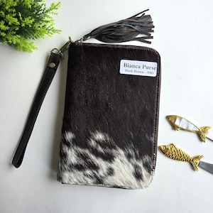 May include: A dark brown and white cowhide Bianca Purse with a wrist strap and a dark brown tassel. The purse has a zip closure and a small white label that reads "Bianca Purse Dark Brown - DB1".