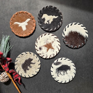 May include: Six round leather coasters with different designs. The coasters are made of cowhide and have a stitched edge. The designs include a longhorn steer, a horse, a star, a bear, and a buffalo.