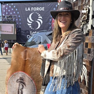 May include: A woman wearing a brown and white cowhide jacket with fringe, and a black hat. She is holding a large cowhide with a logo. The background includes a sign that says "LA FORGE".