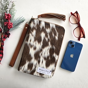 May include: A rectangular light brown and white cowhide Bianca Purse with a brown leather wrist strap and tassel. A blue smartphone and red eyeglasses are next to the purse. The purse has a tag that reads "Bianca Purse Light Brown - L67."