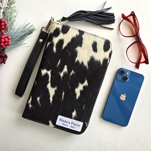 May include: A black and white cowhide purse with a wrist strap and a black tassel. The purse is next to a pair of red glasses and a blue smartphone. The purse has a label that reads "Bianca Purse Black - BK6".