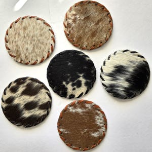 Op de afbeelding: Zes ronde onderzetters van koeienhuid met leren gestikte randen. De onderzetters hebben verschillende natuurlijke vachtpatronen in bruin, zwart en wit. Deze decoratieve onderzetters zijn ontworpen om oppervlakken te beschermen tegen drankringen.
