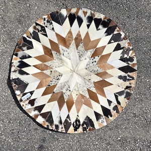 Op de afbeelding: Een rond, veelkleurig koeienhuidkleed met een stervormig patroon. Het kleed is gemaakt van verschillende tinten bruin, wit en zwart.