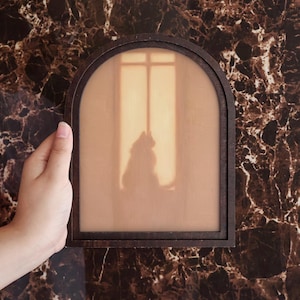 May include: An arched wooden frame containing a translucent panel that displays a silhouette of a cat sitting in front of a window. The frame is dark brown, and the central image features warm, light-toned colors depicting a feline figure.