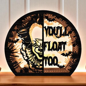 May include: A wooden, semi-circular Halloween decoration featuring a carved image of a clown and the text "YOU'LL FLOAT TOO." The design includes bats and a warm, glowing light from within. The piece is set on a wooden surface.