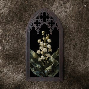 May include: A dark brown wooden wall hanging featuring a pointed gothic arch frame with intricate quatrefoil cutouts at the top. Inside the frame is a vertical rectangular print of white lily of the valley flowers with green leaves on a black background.