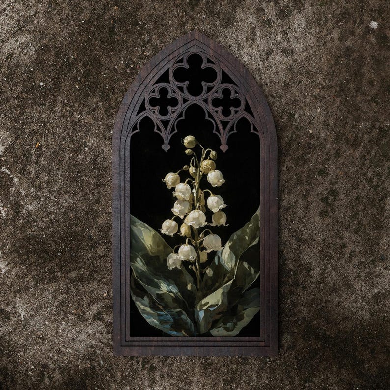 May include: A dark brown wooden wall hanging featuring a pointed gothic arch frame with intricate quatrefoil cutouts at the top. Inside the frame is a vertical rectangular print of white lily of the valley flowers with green leaves on a black background.