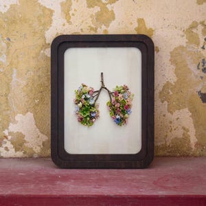 May include: A rectangular dark brown wooden frame containing a light-colored backing. Inside the frame, small twigs and a variety of tiny, colorful flowers are arranged to form the shape of human lungs, featuring pink, blue, and green floral tones.