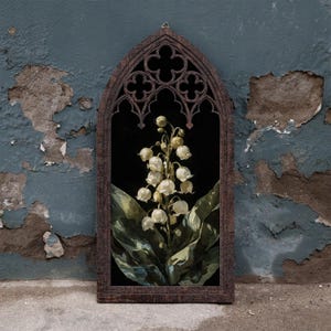 May include: A dark brown wooden wall hanging featuring a pointed gothic arch frame with intricate quatrefoil cutouts at the top. The center displays a detailed illustration of white lily of the valley flowers with green leaves on a black background.