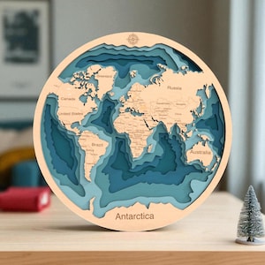 Peut inclure: Une carte du monde en bois de forme circulaire, avec des continents et des océans bleus et beiges en couches. La carte comprend les noms des pays et est montée sur un cadre en bois clair. Le mot "Antarctique" est imprimé en bas.