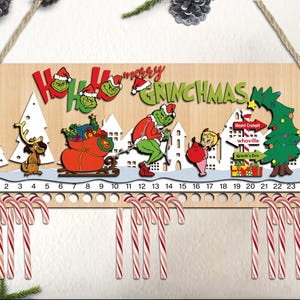 Può includere: Un calendario dell'avvento in legno a tema Grinch. Presenta un personaggio verde Grinch che tira una slitta rossa con i regali. Il calendario ha 24 caselle numerate con supporti per bastoncini di zucchero. Il testo "Ho Ho Ho Merry Grinchmas" è visualizzato nella parte superiore.