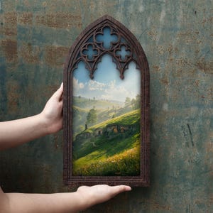 May include: A dark brown wooden frame with a pointed gothic arch top featuring decorative quatrefoil cutouts. The frame encloses a landscape print of rolling green hills, grassy slopes, and small earth-covered dwellings under a blue sky.