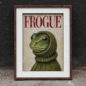 Puede incluir: Una lámina enmarcada que muestra una rana verde con capucha y suéter de punto verde. La palabra FROGUE está impresa en letras mayúsculas rojas grandes en la parte superior. La obra está en un marco de madera marrón oscuro con un paspartú blanco.