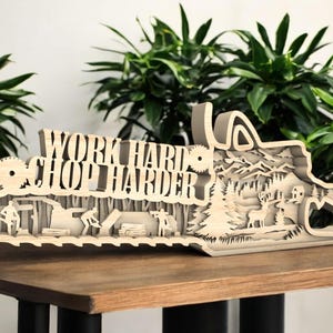 May include: A wooden chainsaw-shaped decorative piece with the text "WORK HARD CHOP HARDER". The design features layered cutouts of lumberjacks, trees, mountains, and wildlife. The piece is displayed on a wooden surface.