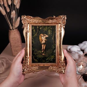 Op de afbeelding: Een rechthoekige fotolijst met een sierlijke, in reliëf gemaakte gouden afwerking met bloemmotieven en krulwerk. De lijst bevat een print van een schilderij met een figuur met lang rood haar in een donkere, dichte bosrijke omgeving.