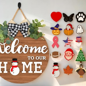 以下が含まれることがあります： 「ようこそ、我が家へ」の文字と雪だるまのイラストが描かれた丸い木製ドアハンガー。ハンガーは黒と白のチェック柄のリボンで飾られています。画像には、ハート、蝶、足跡、クローバー、魔女の帽子、ノーム、幽霊、コウモリ、リボン、星、帽子、カボチャ、フットボール、サンタクロース、クリスマスツリー、雪片、雪だるま、カエデの葉、七面鳥、貝殻など、さまざまな装飾的な木製の切り抜きも含まれています。