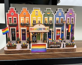 May include: A colorful Pride-themed decorative piece featuring a row of houses painted in rainbow colors. Each house has a door, windows, and a flower box. The sign reads "Chandler's Pride" and "Love is Love". A rainbow flag is displayed.