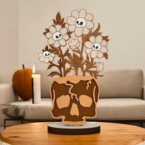May include: A wooden Halloween decoration featuring a skull-shaped vase with flowers. The flowers have skull faces in the center. The vase and flowers are brown and tan. A lit candle and pumpkin are in the background. The decoration is on a round wooden table.