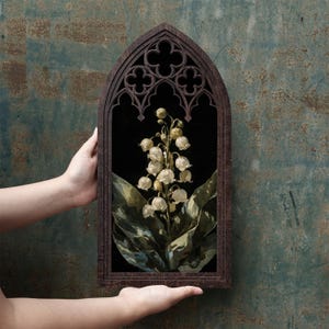 May include: A dark brown wooden frame with a pointed gothic arch top and decorative quatrefoil cutouts. Inside the frame is a botanical illustration of a lily of the valley plant with white bell-shaped flowers and large green leaves on a black background.