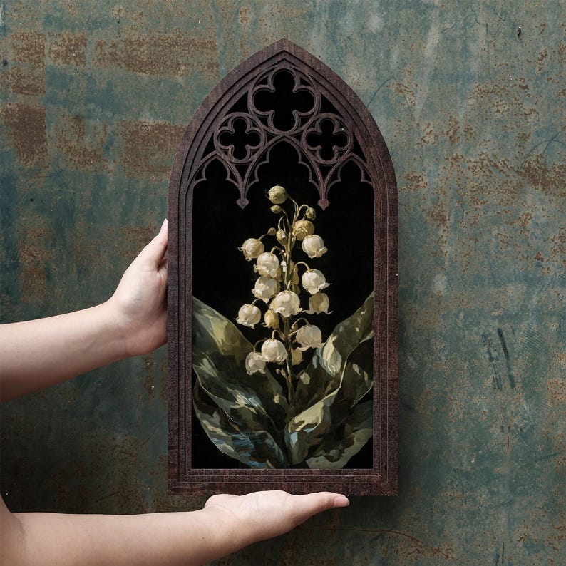 May include: A dark brown wooden frame with a pointed gothic arch top and decorative quatrefoil cutouts. Inside the frame is a botanical illustration of a lily of the valley plant with white bell-shaped flowers and large green leaves on a black background.