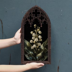 May include: A dark brown wooden frame with a pointed gothic arch top and decorative quatrefoil cutouts. Inside the frame is a botanical illustration of a lily of the valley plant with white bell-shaped flowers and large green leaves on a black background.