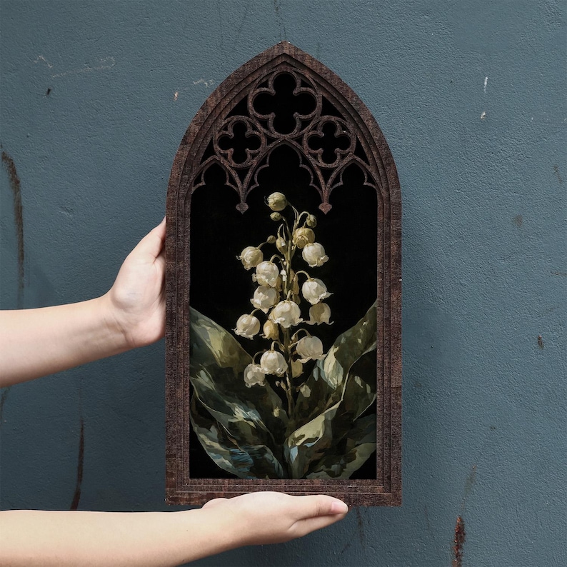 May include: A dark brown wooden frame with a pointed gothic arch top and decorative quatrefoil cutouts. Inside the frame is a botanical illustration of a lily of the valley plant with white bell-shaped flowers and large green leaves on a black background.