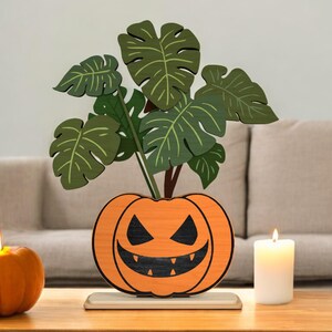 May include: A wooden Halloween decoration featuring a jack-o'-lantern with a carved face, holding green monstera leaves. An orange pumpkin candle and a white candle are on a wooden table.