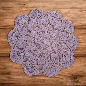 May include: A delicate, crocheted doily in a light purple color. The doily has a circular shape with intricate patterns and a scalloped edge.