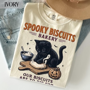May include: An ivory t-shirt featuring a black cat kneading dough on a wooden board with a bowl, a pumpkin, and small ghost figures. The text reads SPOOKY BISCUITS BAKERY and OUR BISCUITS ARE TO DIE FOR in orange and dark purple lettering.