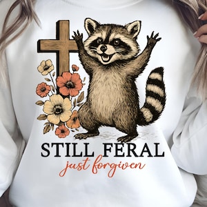 May include: White sweatshirt featuring a raccoon illustration with a cross and flowers. The raccoon has its arms raised. Text on the sweatshirt reads "STILL FERAL just forgiven". The design incorporates brown, orange, and black colors.