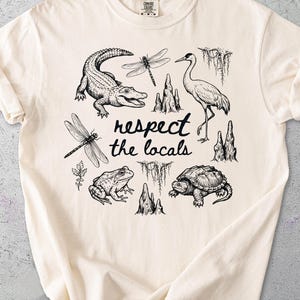 May include: Ivory-colored t-shirt featuring a black and white illustration of various swamp creatures, including an alligator, dragonfly, frog, turtle, and crane. The text "respect the locals" is centered below the illustrations.