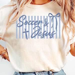 May include: Ivory-colored t-shirt with the text "Soccer & Jesus" in light blue. The text is set against a backdrop of vertical light blue stripes and a bow graphic. The shirt is a casual wear item.