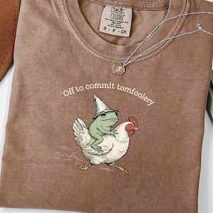 Könnte beinhalten: Espressofarbenes T-Shirt mit Rundhalsausschnitt. Das Shirt zeigt eine skurrile Illustration eines Frosches mit Zaubererhut, der auf einem Huhn reitet, mit dem Text "Off to commit tomfoolery". Eine silberne Halskette mit Anhänger liegt auf dem Shirt.