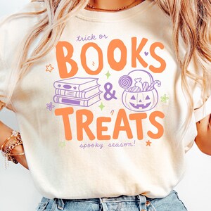 May include: An ivory short-sleeve t-shirt featuring a graphic with the text "trick or BOOKS & TREATS spooky season!" in orange and purple. The design includes a stack of books and a jack-o'-lantern bucket filled with candy and lollipops.