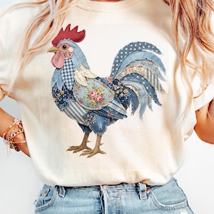 May include: Cream-coloured t-shirt with a rooster design. The rooster is made up of various blue fabric patterns, including floral, polka dot, and striped designs. The rooster has a red comb and wattle. The word "IVORY" is printed at the top left.