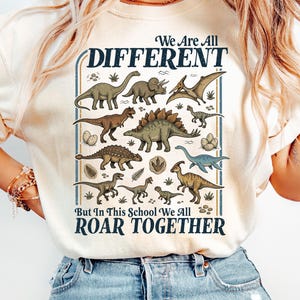 Könnte beinhalten: Elfenbeinfarbenes T-Shirt mit einem Dinosaurier-Motiv. Das Design zeigt verschiedene Dinosaurier-Illustrationen und den Text "We Are All Different But In This School We All Roar Together". Das Shirt besteht aus einem weichen Material.