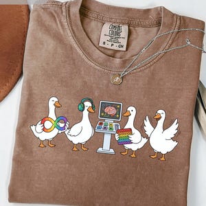 May include: Espresso-colored t-shirt with a graphic of four white ducks. One duck holds a rainbow infinity symbol, another wears headphones, and a third stands near a computer screen. The fourth duck has its wings spread. A silver necklace is also visible.