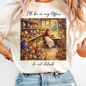 May include: Ivory-colored t-shirt featuring a watercolor illustration of a pottery studio. The image shows a person working at a pottery wheel, surrounded by shelves of pottery. Text on the shirt reads "I'll be in my Office do not disturb."