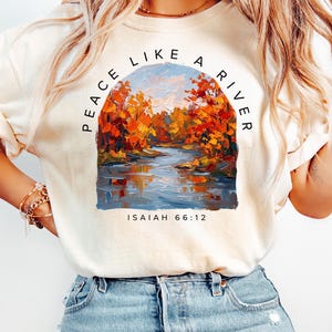 May include: An ivory t-shirt featuring a graphic of an impressionist-style landscape painting of a river surrounded by autumn trees with orange and red foliage. The text PEACE LIKE A RIVER is arched above the image, and ISAIAH 66:12 is below.