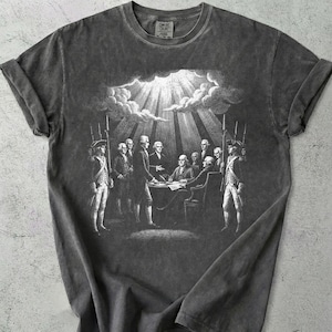 Puede incluir: Camiseta gris oscuro con una ilustración histórica de hombres alrededor de una mesa, custodiados por soldados. La palabra "PEPPER" está impresa en la esquina superior izquierda. La camiseta tiene un aspecto vintage y desgastado.