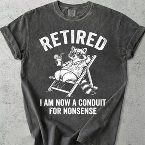 May include: Dark gray t-shirt with the word "RETIRED" in white block letters. A cartoon raccoon wearing sunglasses and a Hawaiian shirt is lounging in a beach chair, holding a drink. The text "! AM NOW A CONDUIT FOR NONSENSE" is below.