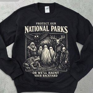 May include: Black sweatshirt with a graphic that reads "Protect Our National Parks" and "Or We'll Haunt Your Backyard." The graphic features a variety of spooky creatures, including a frog, ghosts, and a skeletal deer.
