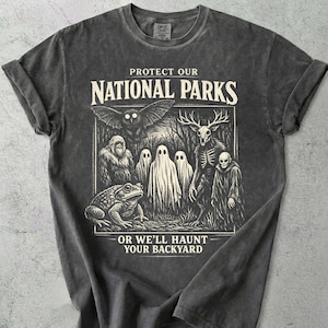 Può includere: T-shirt grigio scuro con maniche arrotolate. La grafica recita "PROTECT OUR NATIONAL PARKS" e "OR WE'LL HAUNT YOUR BACKYARD". Il design include un pipistrello, un bigfoot, fantasmi, un teschio di cervo, una rana e la morte.