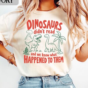 May include: Ivory-colored t-shirt with a red and green dinosaur graphic. The text reads "DINOSAURS didn't read and we know what HAPPENED TO THEM". Features a cartoon volcano, palm tree, and two dinosaurs.