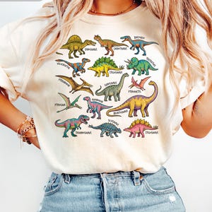 Op de afbeelding: Ivoor kleurig T-shirt met een kleurrijke dinosaurusafbeelding. Het ontwerp bevat verschillende dinosaurus illustraties met hun namen, zoals Tyrannosaurus en Stegosaurus. Het shirt heeft een ronde hals en korte mouwen.